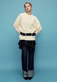 Cremefarbener Oversize-Pullover, schwarzer Gürtel mit zwei großen Schnallen, geschichtete schwarze Rüschenbluse, eng anliegende dunkle Jeans mit ausgefransten Säumen und spitze Stiefel.