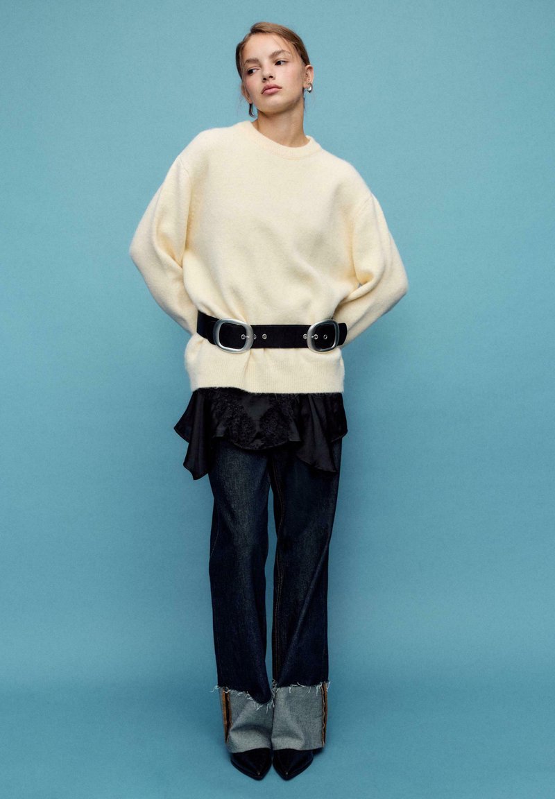 Cremefarbener Oversize-Pullover, schwarzer Gürtel mit zwei großen Schnallen, geschichtete schwarze Rüschenbluse, eng anliegende dunkle Jeans mit ausgefransten Säumen und spitze Stiefel.