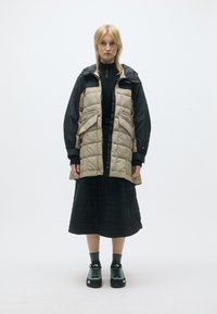 Μπρος, μια ξανθιά γυναίκα φοράει ένα μπεζ επενδυμένο γιλέκο πάνω από ένα μαύρο μπουφάν και φούστα, γκρι κάλτσες και μαύρα αθλητικά παπούτσια.