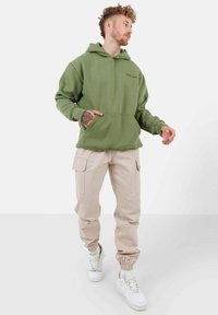 Felpa verde oliva con tasca frontale, abbinata a pantaloni cargo beige con tasche laterali e sneakers bianche con finitura testurizzata.