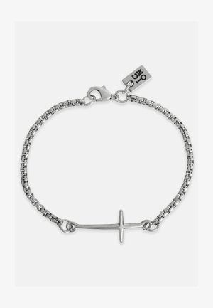Bracelet en chaîne argentée avec un pendentif croix horizontal. Les maillons de la chaîne sont ronds et le fermoir est de type homard avec une étiquette de marque.
