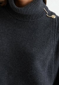 Gros plan sur un pull à col roulé tricoté gris foncé avec une petite broche décorative dorée fixée près du col.