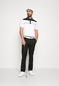Chemise polo blanche avec des rayures noires et jaunes, pantalon noir, ceinture blanche et chaussures de golf blanches. Tenant un club de golf, portant une casquette blanche.