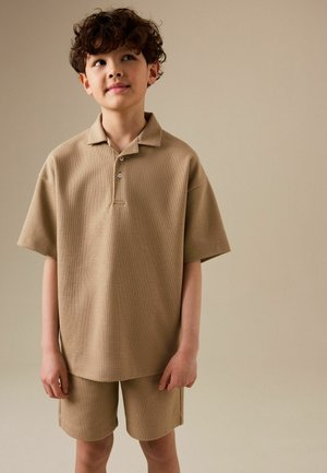 Jongen met krullend haar, gekleed in een beige poloshirt met korte mouwen en bijpassende korte broek, staand tegen een effen beige achtergrond.