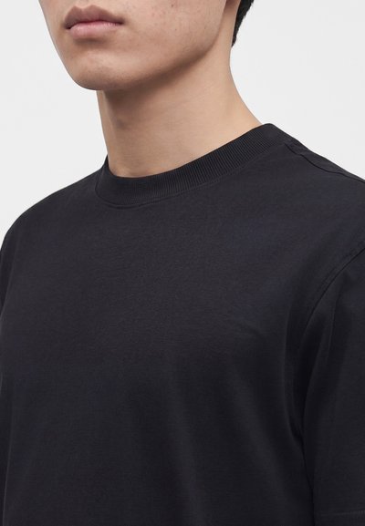 Jeune personne portant un t-shirt noir uni à col rond, montré des épaules jusqu'à juste sous les lèvres sur un fond clair uni.