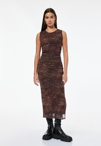 Eine Frau steht in einem ärmellosen, braunen Tiermuster-Midi-Kleid und schwarzen, klobigen Stiefeln vor einem schlichten weißen Hintergrund.