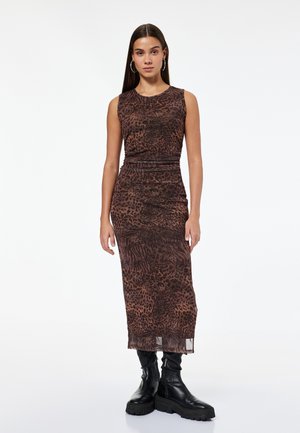 Vrouw die staat in een mouwloze, bruin dierenprint midi-jurk en zwarte chunky laarzen tegen een effen witte achtergrond.