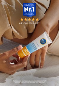 NIVEA Sonnenschutzmittel-Tube mit weißem Körper, blauen und orangefarbenen Akzenten, SPF 50. Eine Hand hält sie vor einem hellen Stoffhintergrund.