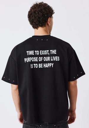 Man met krullend haar die een zwart shirt met witte tekst draagt: "Tijd om te bestaan, het doel van ons leven is gelukkig zijn."