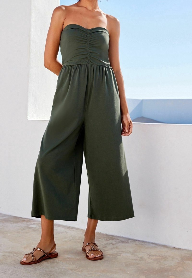 Femme portant une combinaison bustier olive à jambes larges et des sandales brunes tressées, debout devant un mur blanc avec un ciel bleu en arrière-plan.