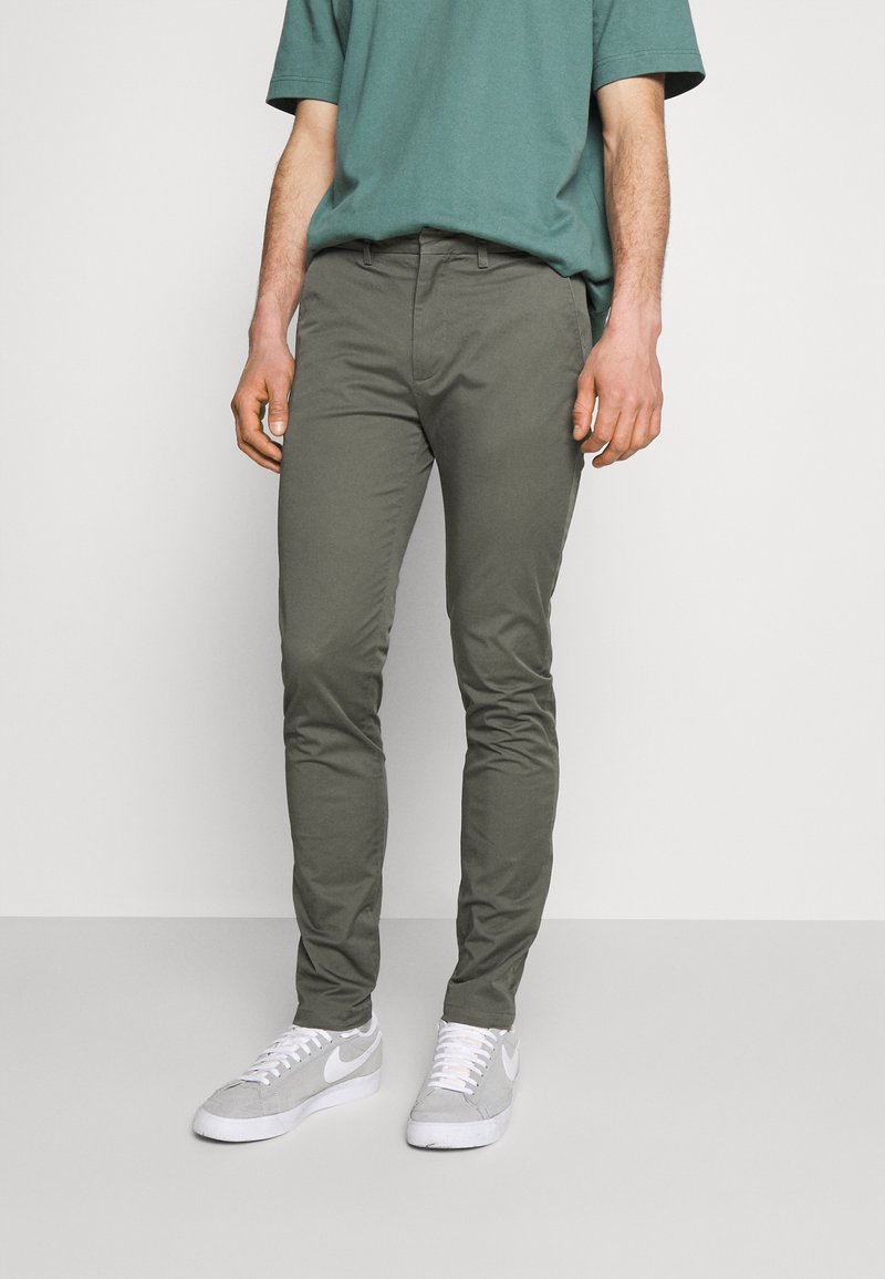 Homme portant un pantalon slim olive, une chemise verte à manches courtes et des baskets Nike gris clair, debout sur un fond uni.