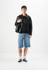 Tânăr stând în picioare, îmbrăcat cu o geacă de piele neagră, un pulover negru, pantaloni scurți din denim albastru și adidași negri, ținând o geantă de piele neagră pe umăr.