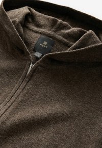 Veste zippée en laine marron avec capuche, montrant l’étiquette intérieure du col avec le nom de la marque "NEXT" et la taille moyenne.