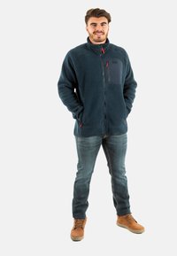 Giacca in pile blu con colletto alto e zip, dotata di tasca sul petto e dettagli rossi. Indossata con jeans in denim e stivali marroni.