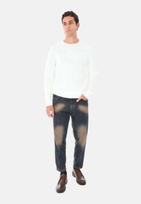 Maglione bianco a maglia, jeans scuri con leggere abrasioni e scarpe marroni con lacci. Il modello si trova in posizione con una mano sui fianchi contro uno sfondo neutro.