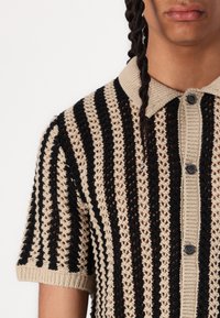 Beige en zwarte gestreepte gebreide poloshirt met korte mouwen, open haakstructuur en knoopsluiting aan de voorkant.