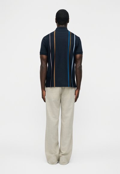 Homme debout de dos, portant un polo bleu marine à rayures verticales multicolores et un pantalon large beige sur un fond uni.