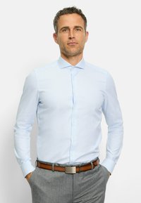 Hellblaues, knöpfbares Hemd mit einem dezent geometrischen Muster, langen Ärmeln und einem Kragen, kombiniert mit grauen Hosen und einem braunen Gürtel.
