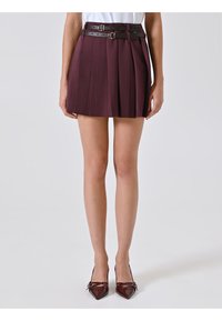 Mini gonna plissettata bordeaux con un design a strati, caratterizzata da due cinture in pelle nera con fibbie argentate. Un paio di tacchi a punta.
