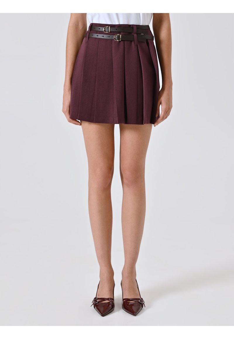 Mini gonna plissettata bordeaux con un design a strati, caratterizzata da due cinture in pelle nera con fibbie argentate. Un paio di tacchi a punta.