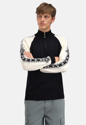 Ung mann med kort hår iført en svart-hvitt halvglidelåssweater med snøfnuggmønster på ermene, stående med armene i kors.