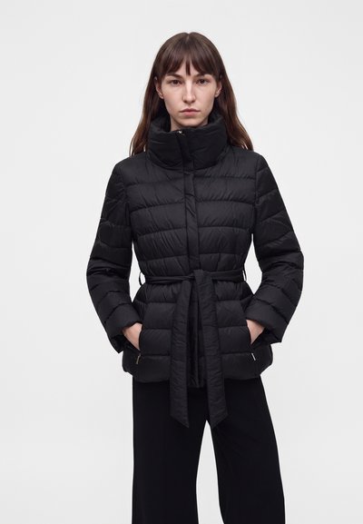 Femme portant une doudoune noire ceinturée avec un col haut et les mains dans les poches, debout devant un fond clair uni.