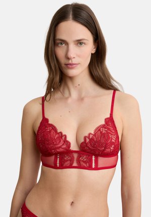 Vrouw met lang bruin haar die een rode kanten beha draagt met bloemenborduursel, dunne bandjes en mesh details, tegen een neutrale achtergrond.