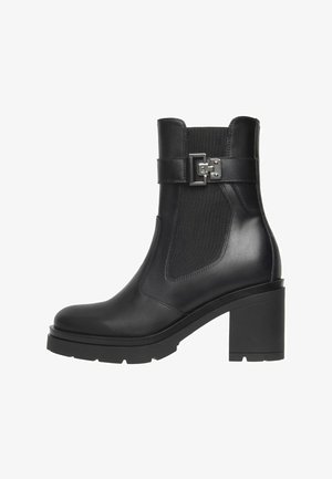 Bottine en cuir noir avec un talon épais, panneau latéral élastique et une sangle décorative en métal argenté sur fond blanc.