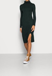 Robe à col roulé vert forêt à manches longues avec une fente latérale, en tissu doux. Associée à des baskets montantes beige.