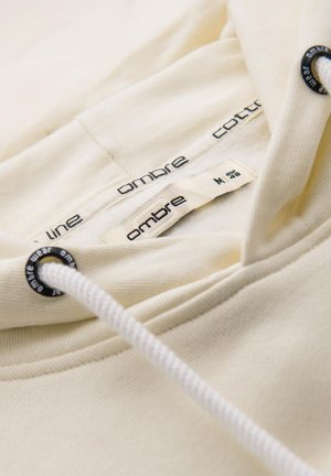 Sweatshirt à capuche en coton crème, avec un cordon de serrage et des étiquettes de marque à l'intérieur. La texture est lisse avec une finition intérieure douce.