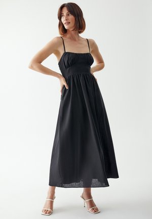 Black maxi dress with thin straps, gathered bodice, and flowy bottom. Smooth texture and minimalist design. Worn with white heeled sandals.