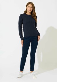 Suéter de punto de cable en color navy con puños de canalé, combinado con jeans oscuros de corte ajustado. El modelo lleva zapatillas blancas, mostrando un atuendo casual.