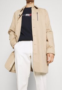 Cappotto beige leggero con chiusura a bottoni, tasca sul petto con cerniera e maniche a risvolto, indossato sopra una t-shirt blu scuro e pantaloni bianchi.