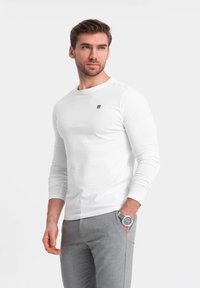Witte langemouw t-shirt met ronde hals en klein logo op de borst, gecombineerd met lichtgrijze broek en een zilveren horloge.
