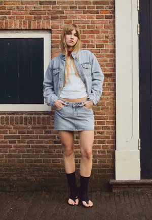 Fiatal nő világos farmerdzsekiben és szoknyában, fehér felsőben, fekete lábszárvédőkkel és papucsban áll egy téglafal előtt, kezét zsebre téve.