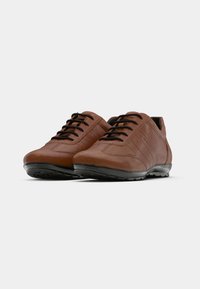 Paire de chaussures décontractées en cuir marron à lacets avec semelles noires, présentée sur un fond gris uni.