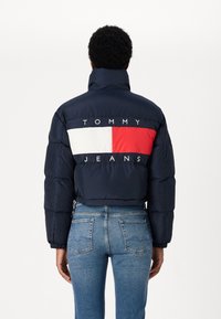 Jūras spēku pufīša jaka ar augstu apkakli, kurai ir horizontāla sarkana un balta svītra ar "TOMMY JEANS" logotipu uz muguras. Tekstūras apdare.
