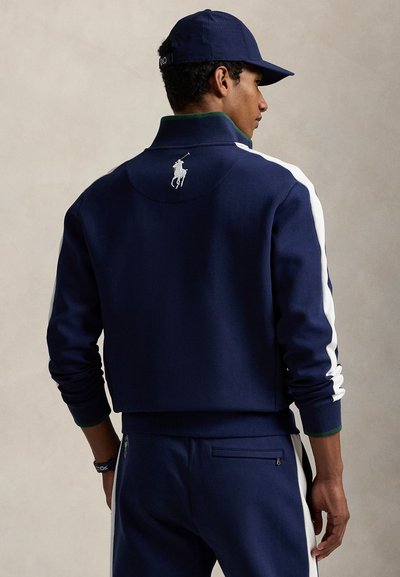 Tmavomodrá športová bunda s bielymi pruhmi na bokoch a zeleným golierom. Na zadnej strane je vyšívané logo a manžety sú rebrované. Kombinovaná s rovnakými nohavicami.