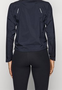 Mörkblå lättviktig jacka med långa ärmar, som har reflektordetaljer och en zippered nederkant. Slät textur, designad för aktiv användning.