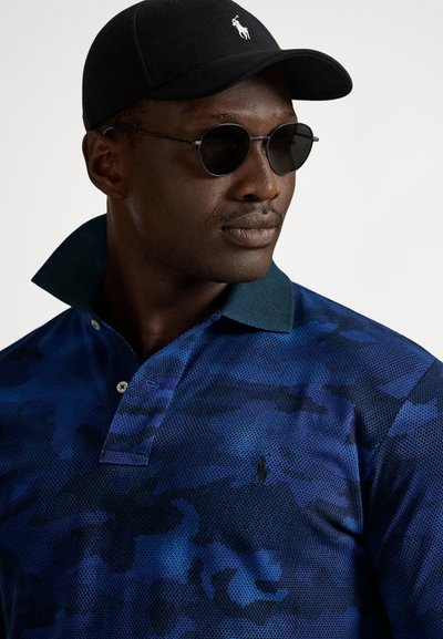Mężczyzna w czarnej czapce, okrągłych ciemnych okularach przeciwsłonecznych i niebieskiej koszuli w kamuflażowy wzór z kołnierzykiem oraz logo na piersi, patrzący w prawo.