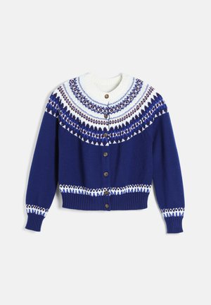 Jeune femme portant un cardigan bleu marine à motif, un top blanc, un pantalon large crème, une ceinture noire, des bottes noires et un sac à épaule noir.