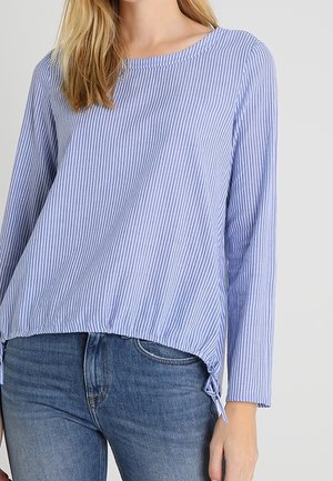 Vrouw draagt een lichtblauw-wit verticale gestreepte blouse met lange mouwen en zijstrikjes, gecombineerd met blauwe spijkerbroeken.