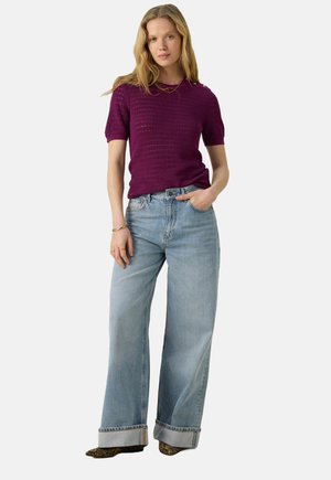 Haut en tricot texturé violet à manches courtes associé à un jean large bleu clair, avec des poignets retroussés et des boutons décoratifs à l'épaule.