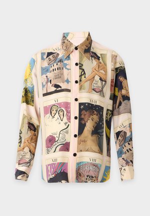 Homme portant une chemise à manches longues imprimée de cartes de tarot, pantalon beige à carreaux, chaussures noires et collier chaîne en argent, debout les mains dans les poches.