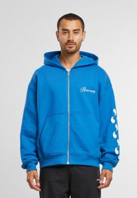 Felpa con zip blu realizzata in un misto cotone, dotata di tasca frontale e dettaglio del logo bianco sul petto, con accenti stilizzati sulle maniche.