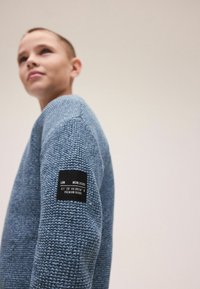 Niebieski sweter z fakturowanym splotem i okrągłym dekoltem. Na rękawie znajduje się czarna etykieta z koordynatami i napisem.