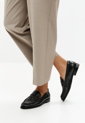 Persoon draagt beige broek en zwarte leren loafers, staande op een witte vloer, één voet licht opgetild.