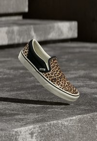 Chaussure Vans sans lacets avec dessus imprimé léopard, côtés noirs, semelle blanche, flottant au-dessus d'escaliers en béton.