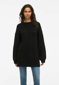 Suéter preto de malha com um corte solto, decote redondo e mangas longas e largas, combinado com jeans azul claro. Tecido texturizado, comprimento até a metade da coxa.