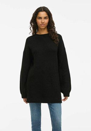 Schwarzer Strickpullover mit lockerer Passform, rundem Halsausschnitt und langen, weiten Ärmeln, kombiniert mit hellblauen Jeans. Strukturiertes Material, midi Länge bis zur Mitte des Oberschenkels.